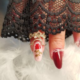 Usługi kosmetyczne Anita Niełacna - Kobieca dłoń z pierścionkami i zdobieniem paznokci: różowy lakier z motywem róży i burgundowy, otoczone piórami i czarną koronką. Elegancki manicure.