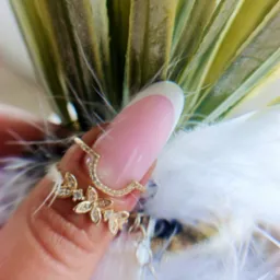 Powiększenie dłoni z delikatnym, różowym manicure french i złotą biżuterią na tle piór i roślin. Subtelne zdobienie paznokci.