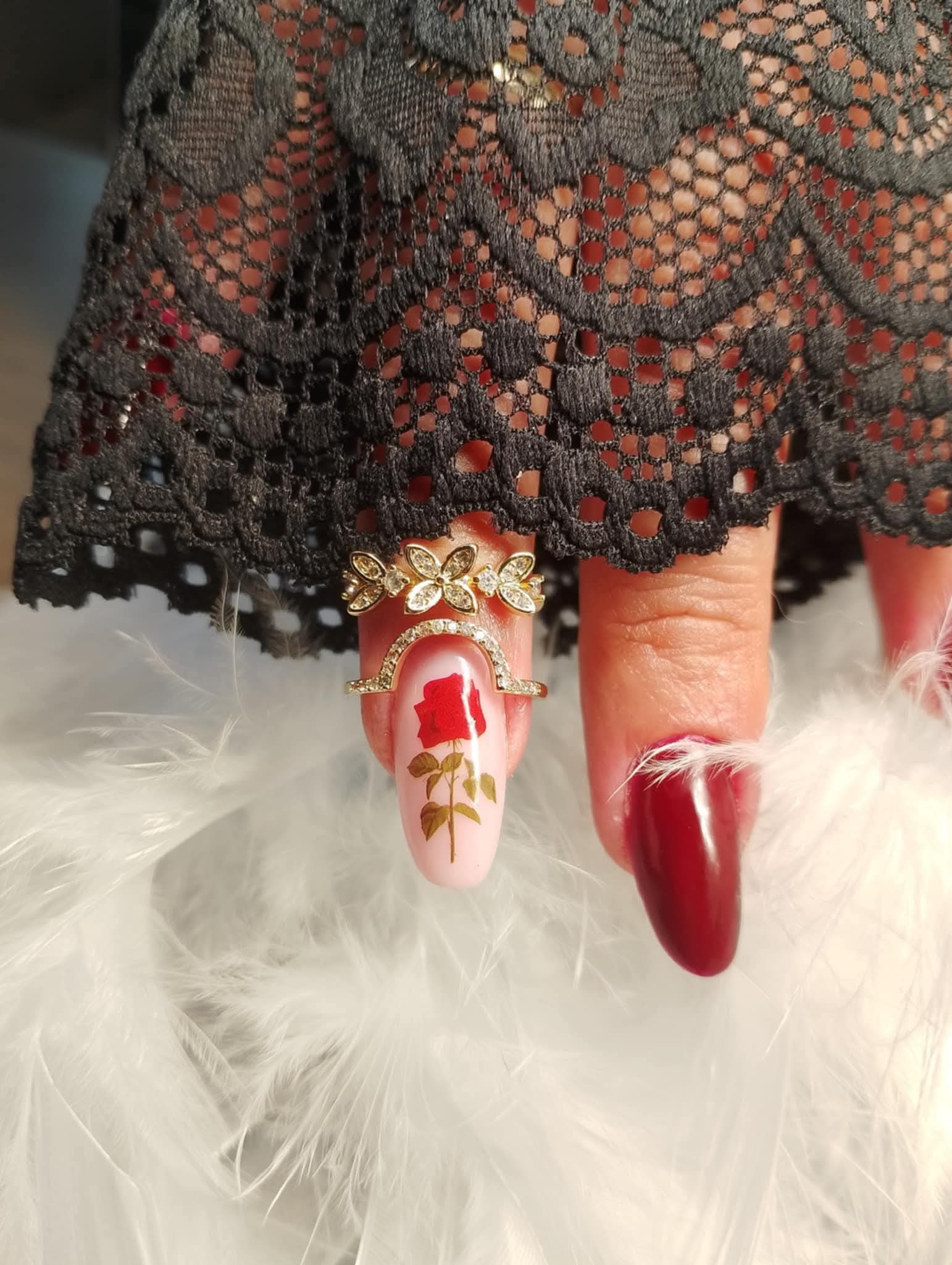 Kobieca dłoń z pierścionkami i zdobieniem paznokci: różowy lakier z motywem róży i burgundowy, otoczone piórami i czarną koronką. Elegancki manicure.