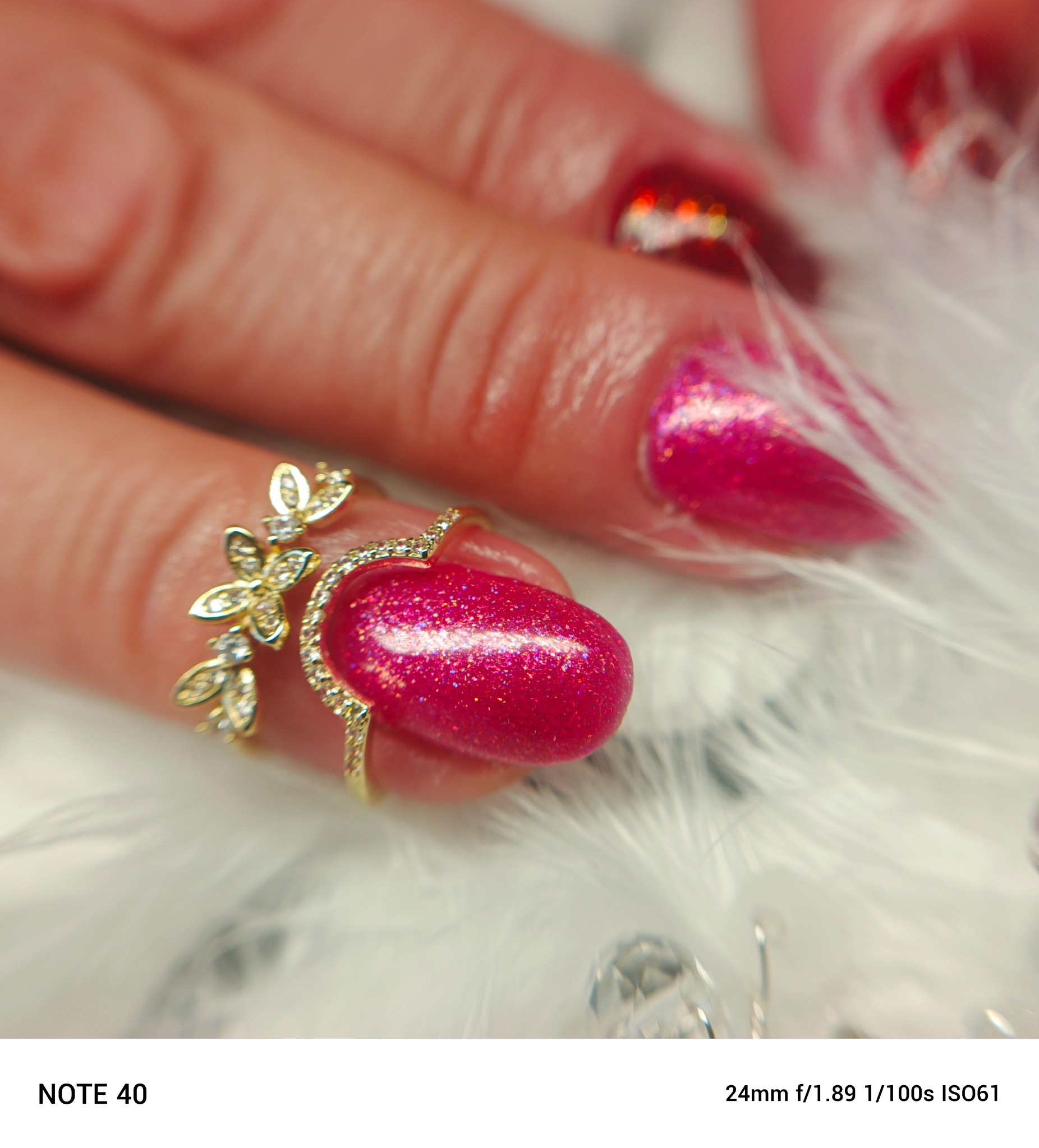 Błyszczący, różowy manicure hybrydowy z efektem brokatu, ozdobiony złotym pierścionkiem z motywem kwiatowym na tle z białych piórek.