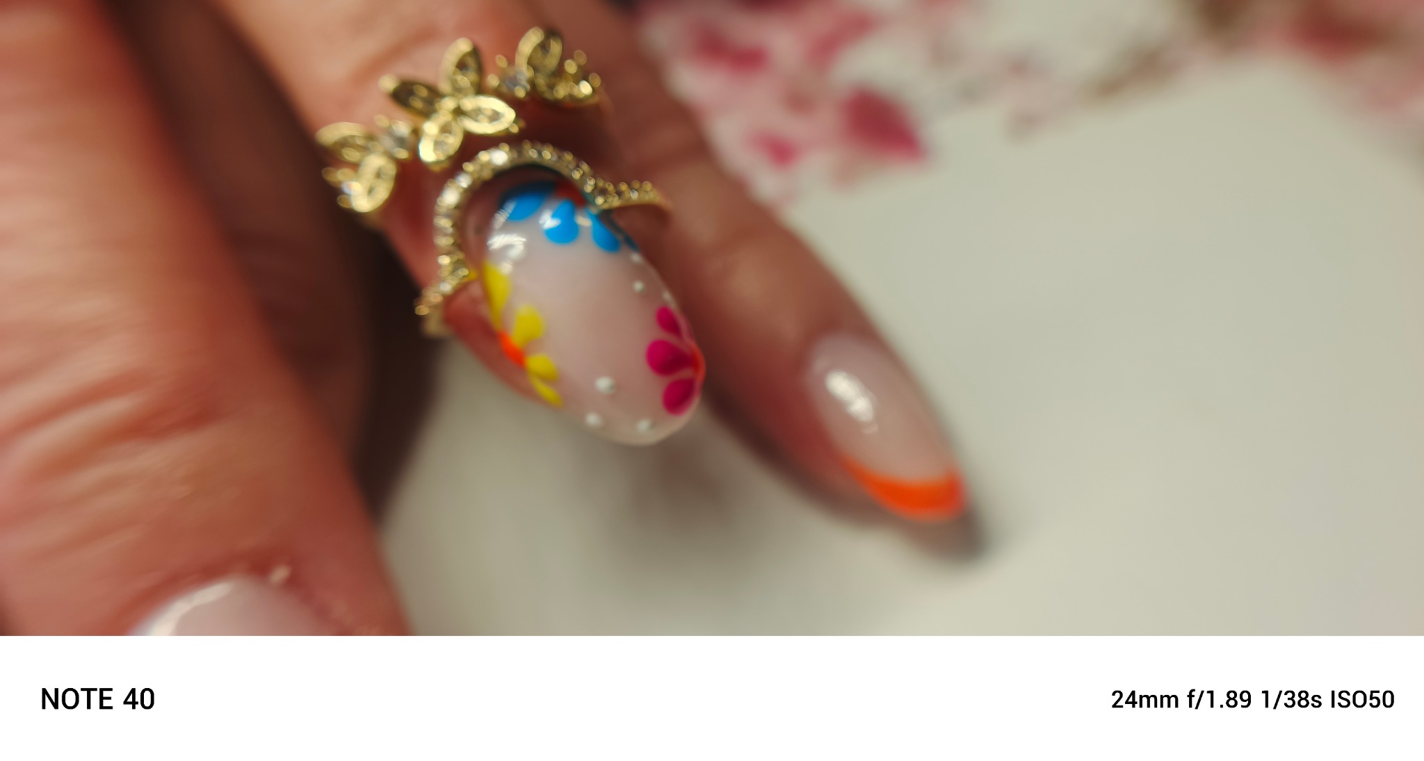 Dłoń z pierścionkiem i zdobionym paznokciem z motywem kwiatowym w kolorach: różowy, żółty i niebieski. Na drugim paznokciu pomarańczowy french manicure.