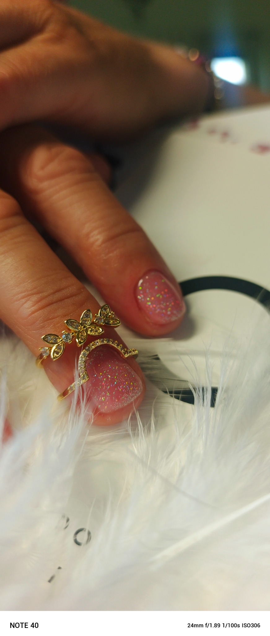 Ręka z różowym manicure z brokatem, ozdobiona złotym pierścionkiem na palcu wskazującym, na tle białych piór. Delikatne, błyszczące wykończenie.