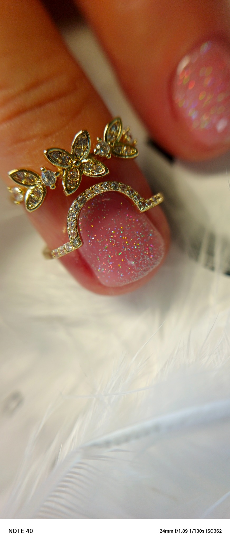 Błyszczący, różowy manicure z brokatem, ozdobiony złotym pierścionkiem w kształcie motyli, na tle białych piór. Efektowny detalizacja i blask.