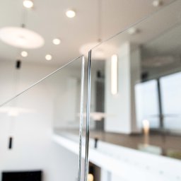 PEKA WOODCRAFT SPÓŁKA Z OGRANICZONĄ ODPOWIEDZIALNOŚCIĄ - Szklana balustrada schodowa z metalowymi mocowaniami, widok z bliska na łączenie tafli szkła, nowoczesne oświetlenie w tle.