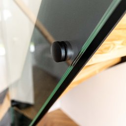 PEKA WOODCRAFT SPÓŁKA Z OGRANICZONĄ ODPOWIEDZIALNOŚCIĄ - Detale szklanej balustrady schodów z drewnianymi stopniami, mocowanej czarnym, okrągłym uchwytem. Widoczne refleksy światła na szkle.