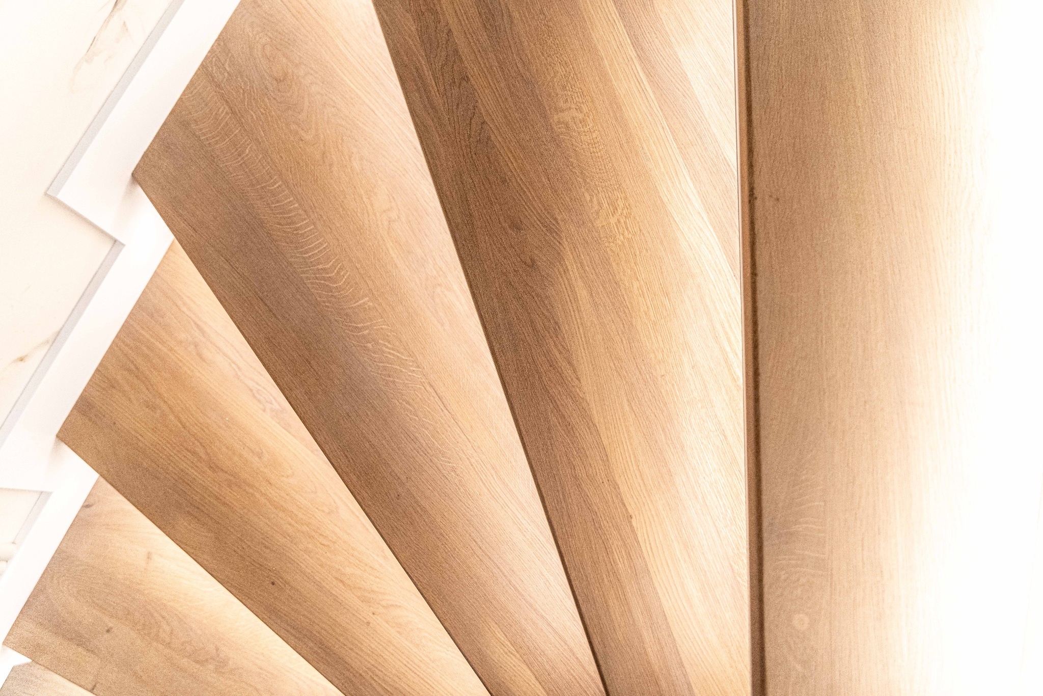 Jasne, drewniane schody spiralne z białymi elementami wykończeniowymi, widok z góry, ukazujący naturalne usłojenie drewna i subtelne oświetlenie.