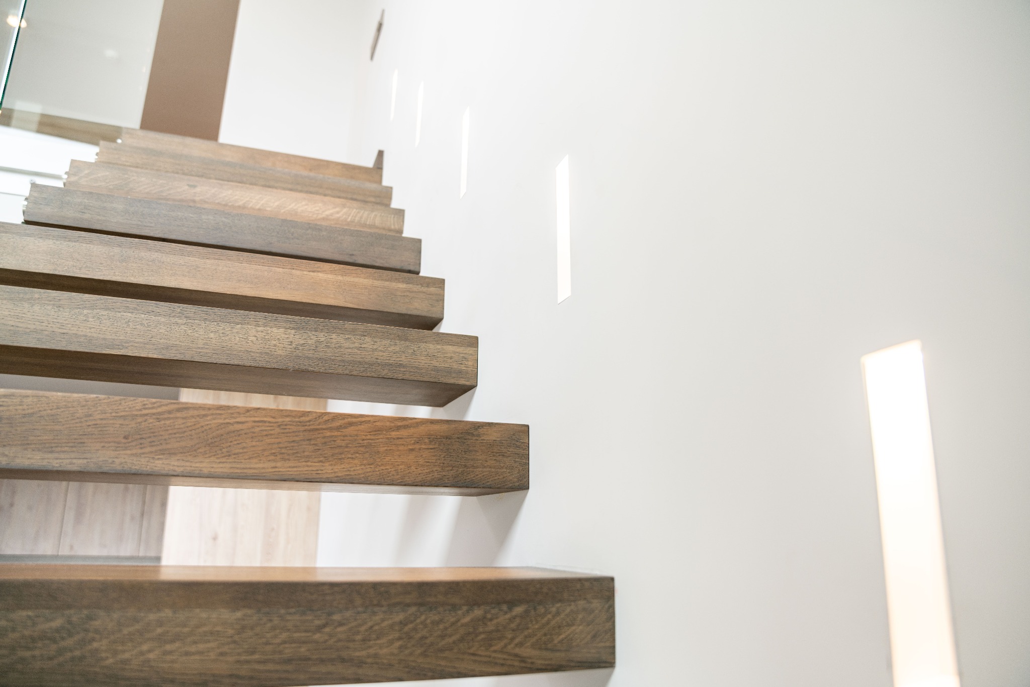 Nowoczesne, drewniane schody wspornikowe z oświetleniem LED w ścianie. Minimalistyczny design z ciepłym odcieniem drewna i białym tłem.