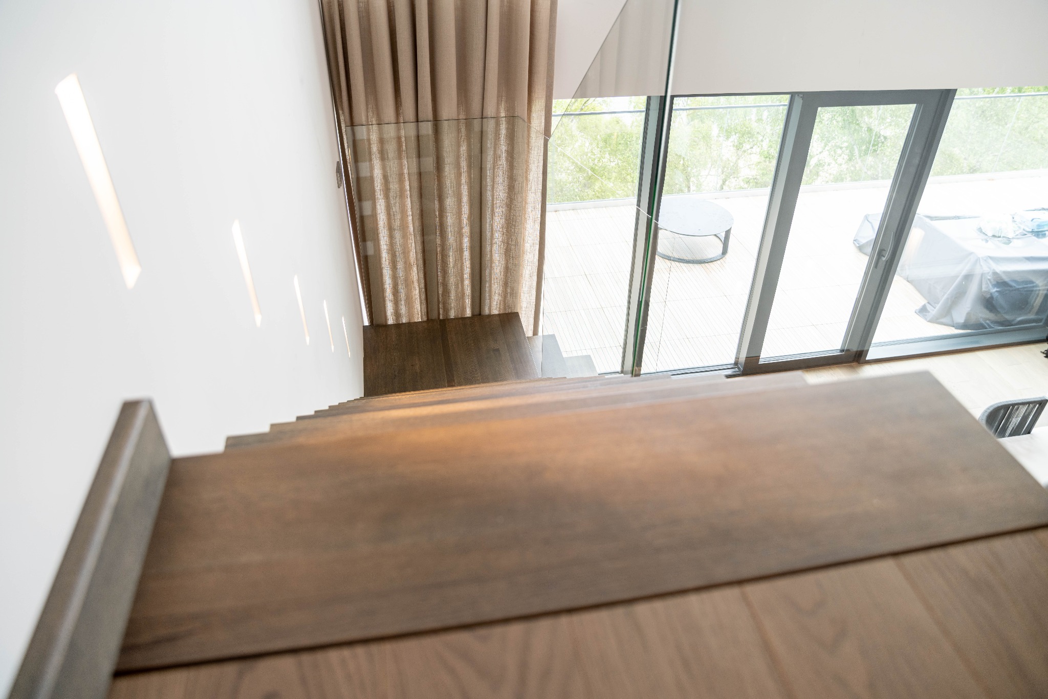 Nowoczesne schody drewniane z ciemnego drewna, widok z góry, z przeszkloną balustradą i widokiem na taras. Minimalistyczny design z elementami naturalnego światła.