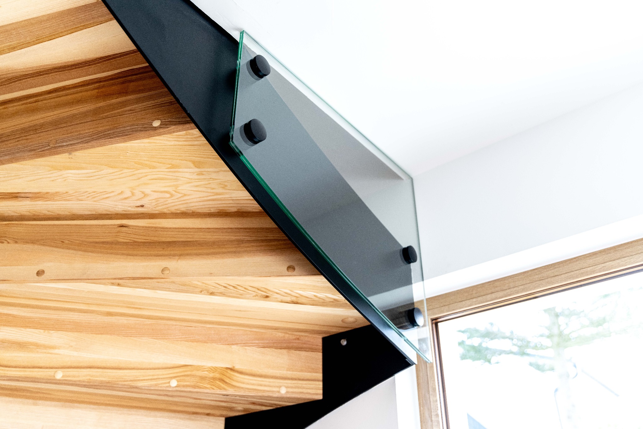 Detal drewnianych schodów z czarną metalową konstrukcją i szklaną balustradą, widok z dołu, jasne wnętrze.