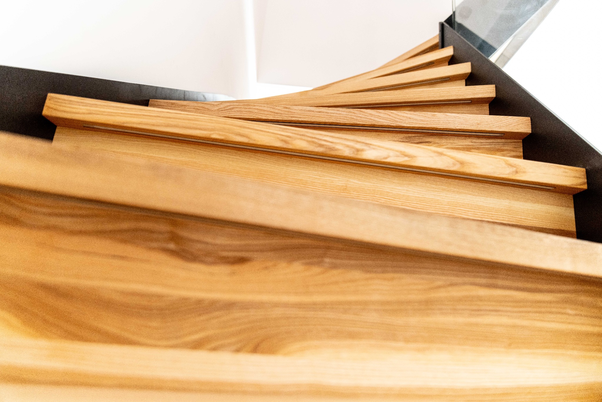 Spiralne schody z jasnego drewna, widok z góry, z czarną barierką i szklanym panelem. Naturalne oświetlenie podkreśla strukturę drewna.