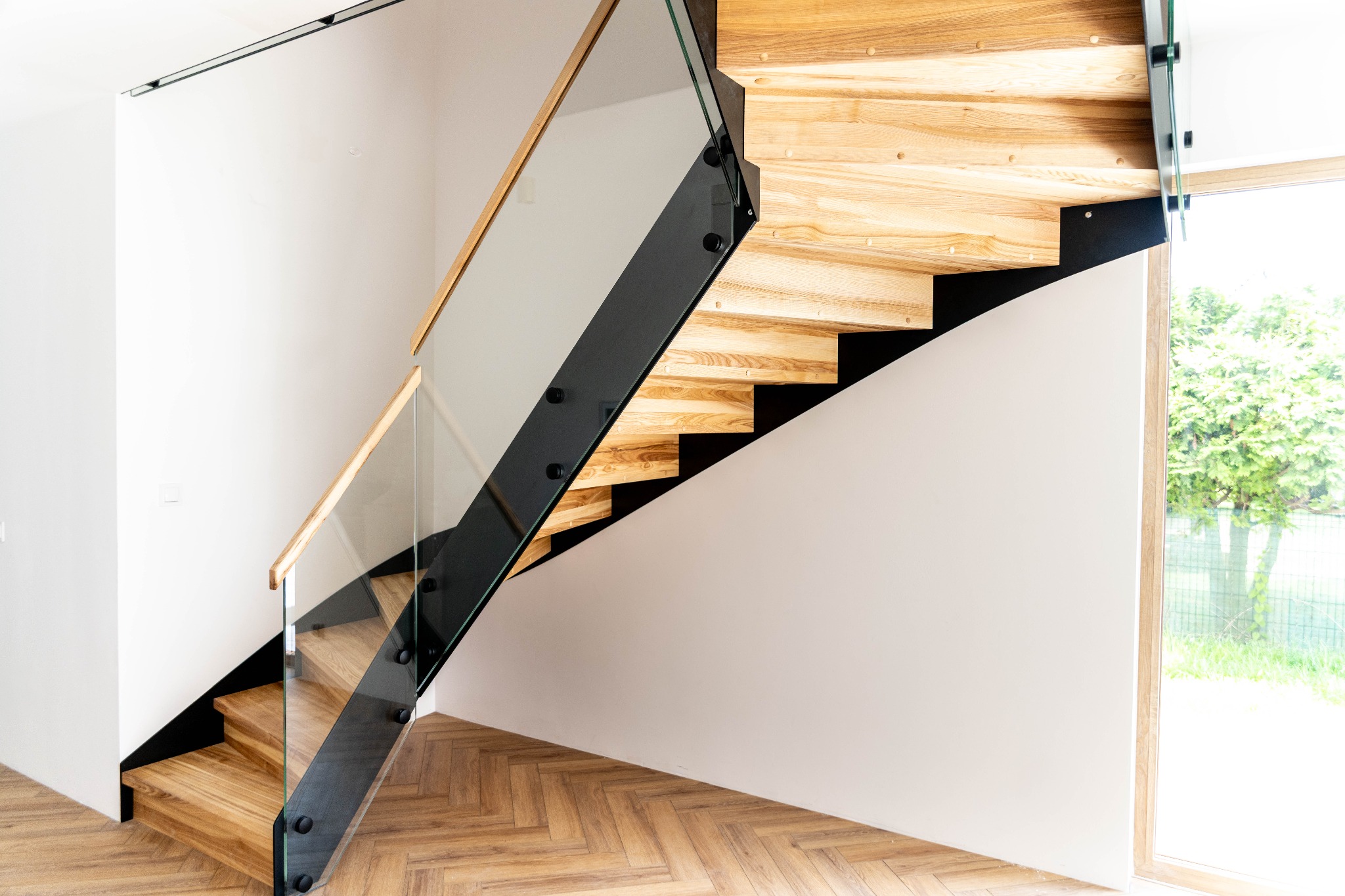 Nowoczesne schody drewniane z czarnymi elementami metalowymi i szklaną balustradą, widok z dołu, parkiet w jodełkę, okno z widokiem na zieleń.