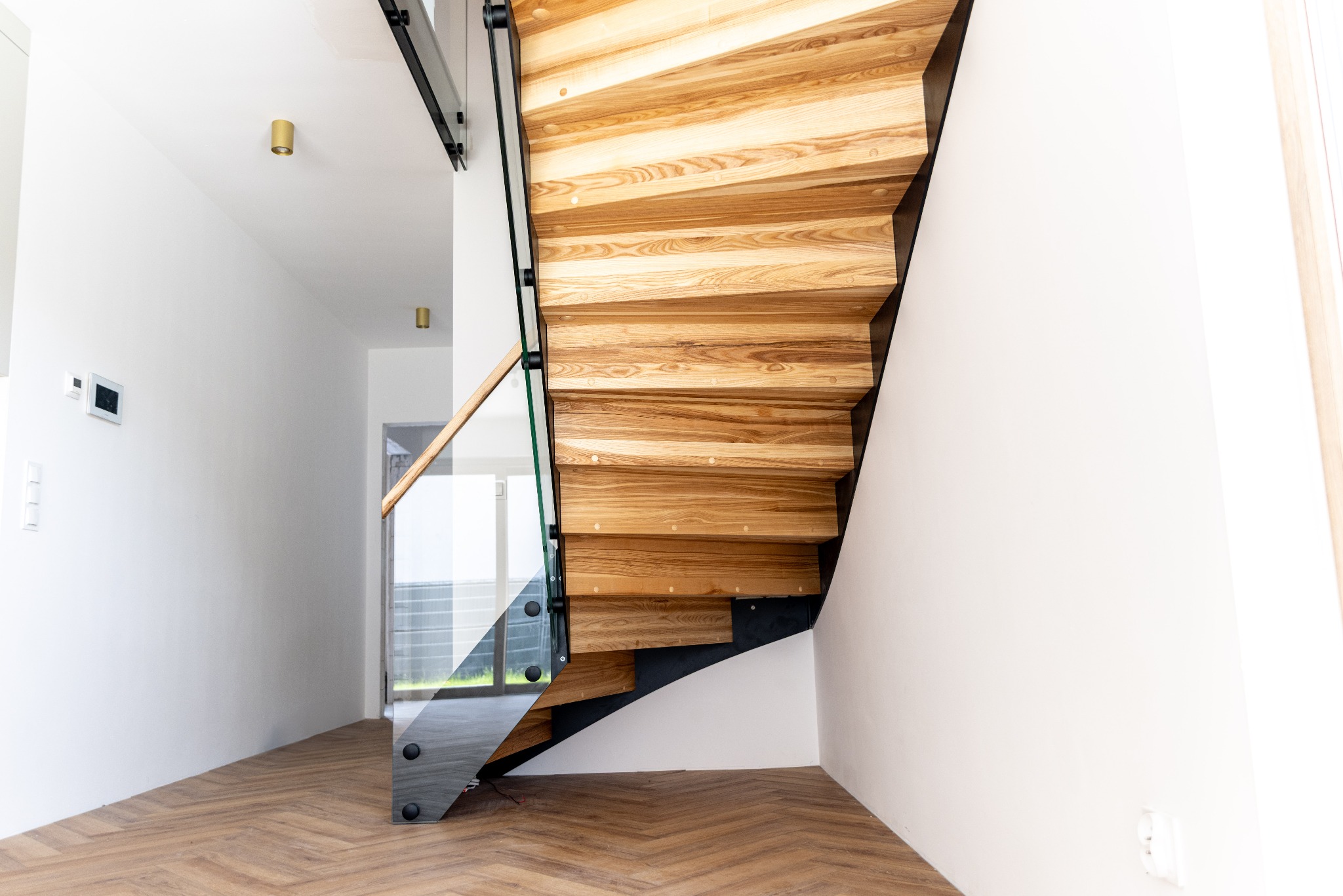 Nowoczesne schody drewniane z elementami metalowymi i szklaną balustradą, zamontowane w jasnym wnętrzu domu w Nowogardzie. Widok z dołu, parkiet w jodełkę.