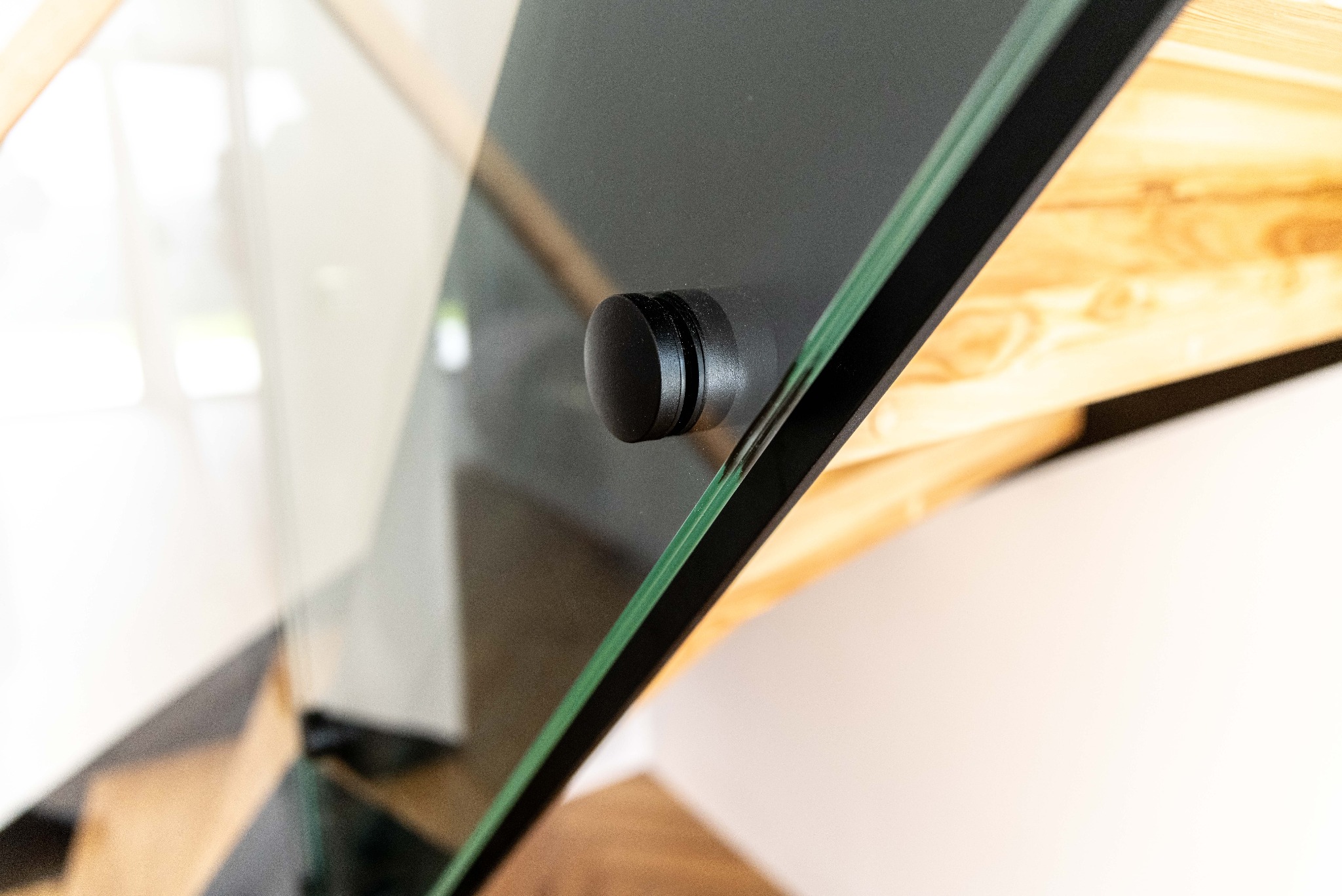 Detale szklanej balustrady schodów z drewnianymi stopniami, mocowanej czarnym, okrągłym uchwytem. Widoczne refleksy światła na szkle.