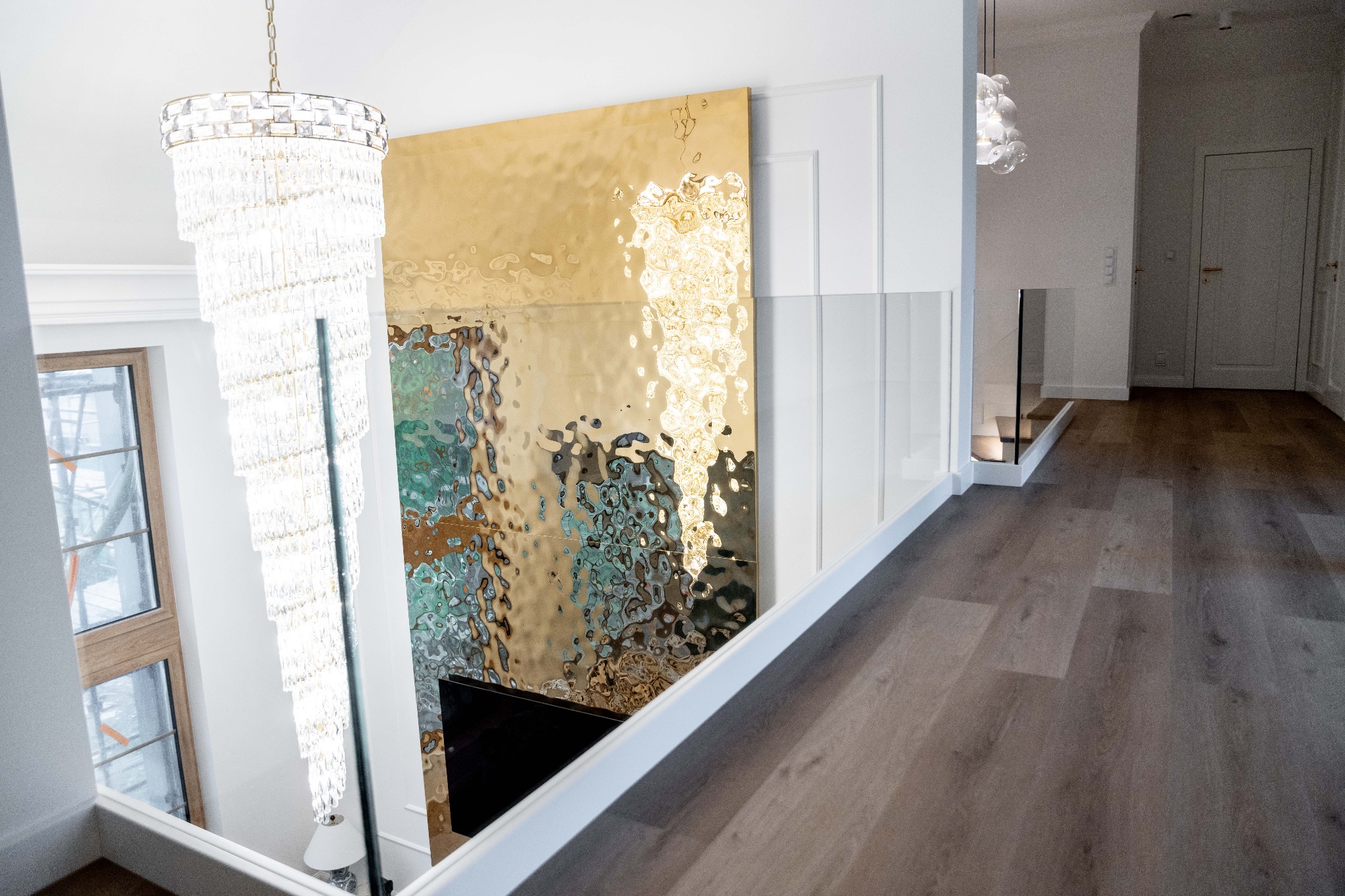 Nowoczesne schody z drewnianymi stopniami i szklaną balustradą, widok z góry. W tle złoty panel dekoracyjny i kryształowy żyrandol.