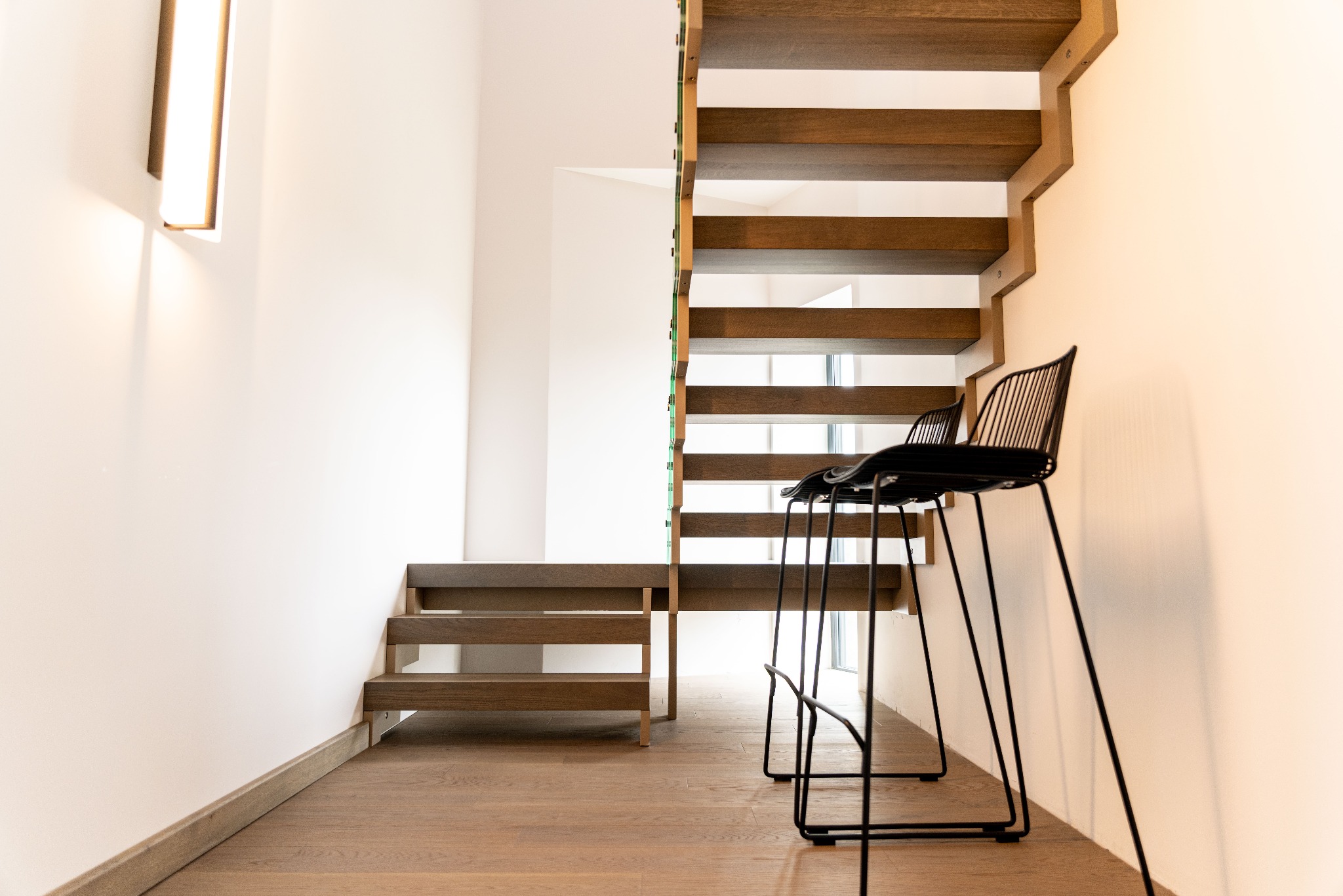 Nowoczesne schody drewniane z ażurową konstrukcją, oświetleniem LED i dwoma krzesłami barowymi w minimalistycznym wnętrzu z drewnianą podłogą.