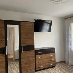 K&V - Sypialnia z drewnianą szafą z lustrem, komodą i telewizorem na ścianie. Jasne ściany, okno z koronkową firanką i drewniana podłoga. Widok na wnętrze przez lustro.