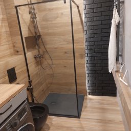 K & V - Nowoczesna łazienka z kabiną walk-in o czarnej ramie, drewnopodobnymi płytkami na ścianach i podłodze. Ściana z ciemnej cegły z oświetleniem LED, czarna toaleta i pralka pod drewnianym blatem.