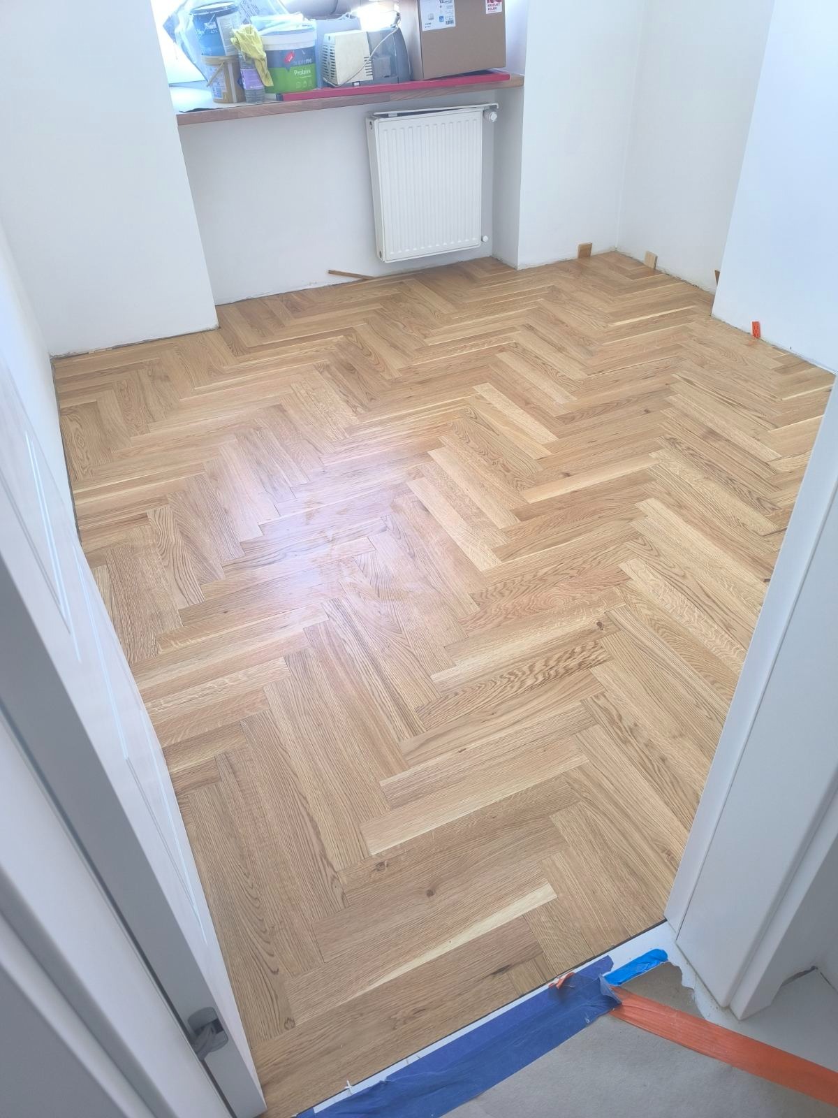 Parkiet ułożony w jodełkę w jasnym pomieszczeniu, widok z góry. Przy oknie farby i akcesoria malarskie. Grzejnik na ścianie, widoczny fragment drzwi.