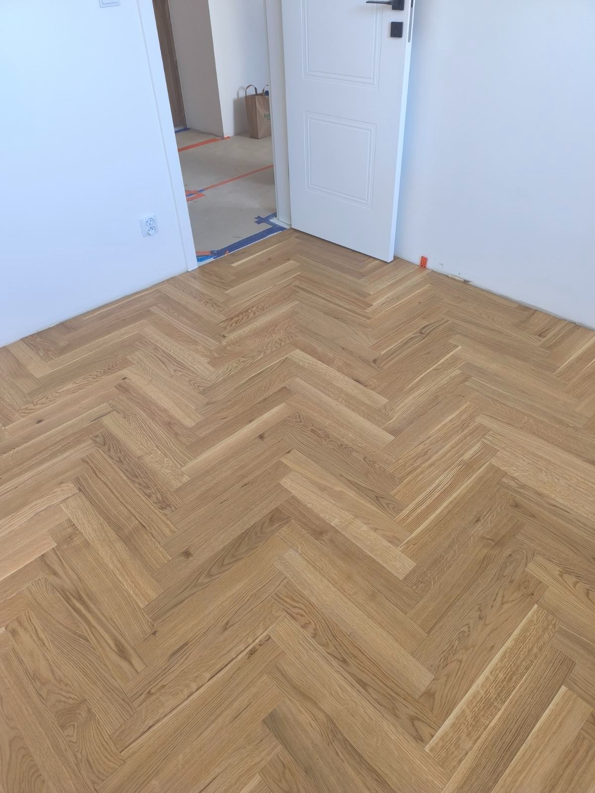 Ukończona podłoga z parkietu ułożonego w jodełkę, widok na białe ściany i otwarte drzwi do kolejnego pomieszczenia w trakcie prac wykończeniowych.