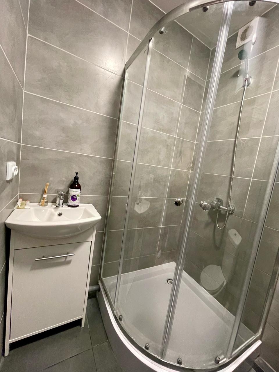 Nowoczesna łazienka z kabiną prysznicową narożną i białą szafką z umywalką. Szare płytki na ścianach i podłodze. Minimalistyczny design.