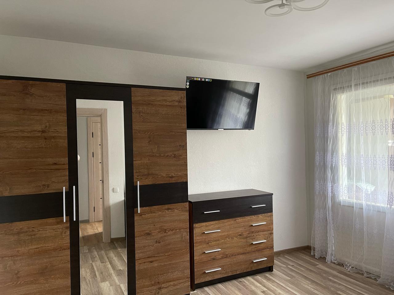 Sypialnia z drewnianą szafą z lustrem, komodą i telewizorem na ścianie. Jasne ściany, okno z koronkową firanką i drewniana podłoga. Widok na wnętrze przez lustro.