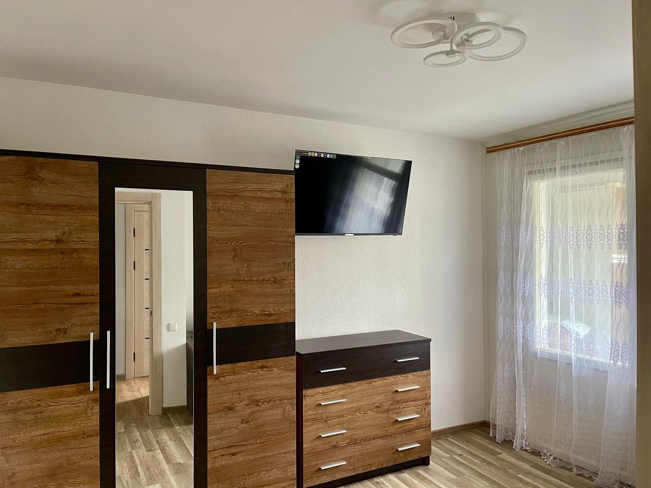 Nowoczesna sypialnia: drewniana szafa z lustrem, komoda, telewizor na ścianie, okno z firanką i designerska lampa sufitowa. Jasne wnętrze z elementami drewna.