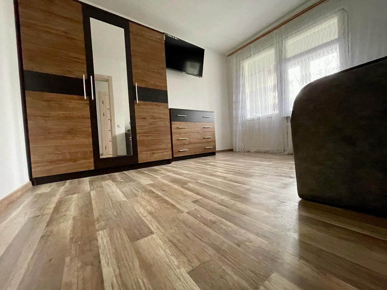 Jasny pokój z drewnianą podłogą, szafą z lustrem, komodą i telewizorem na ścianie. Widok z niskiej perspektywy, podkreślający przestronność wnętrza.