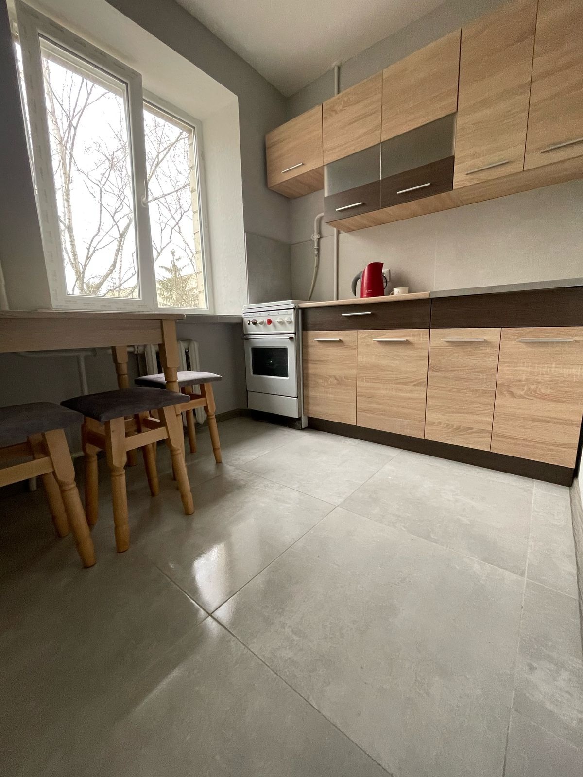 Jasna kuchnia po remoncie: szare płytki podłogowe, drewniane szafki wiszące i stojące, okno z widokiem na drzewa. Minimalistyczny design.