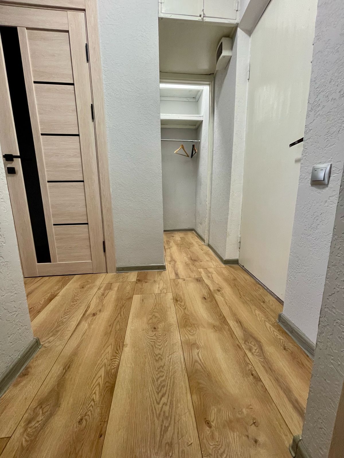 Nowoczesny korytarz z panelami podłogowymi imitującymi drewno, widok na garderobę z wieszakami. Drzwi w kolorze jasnego drewna z czarnymi akcentami.