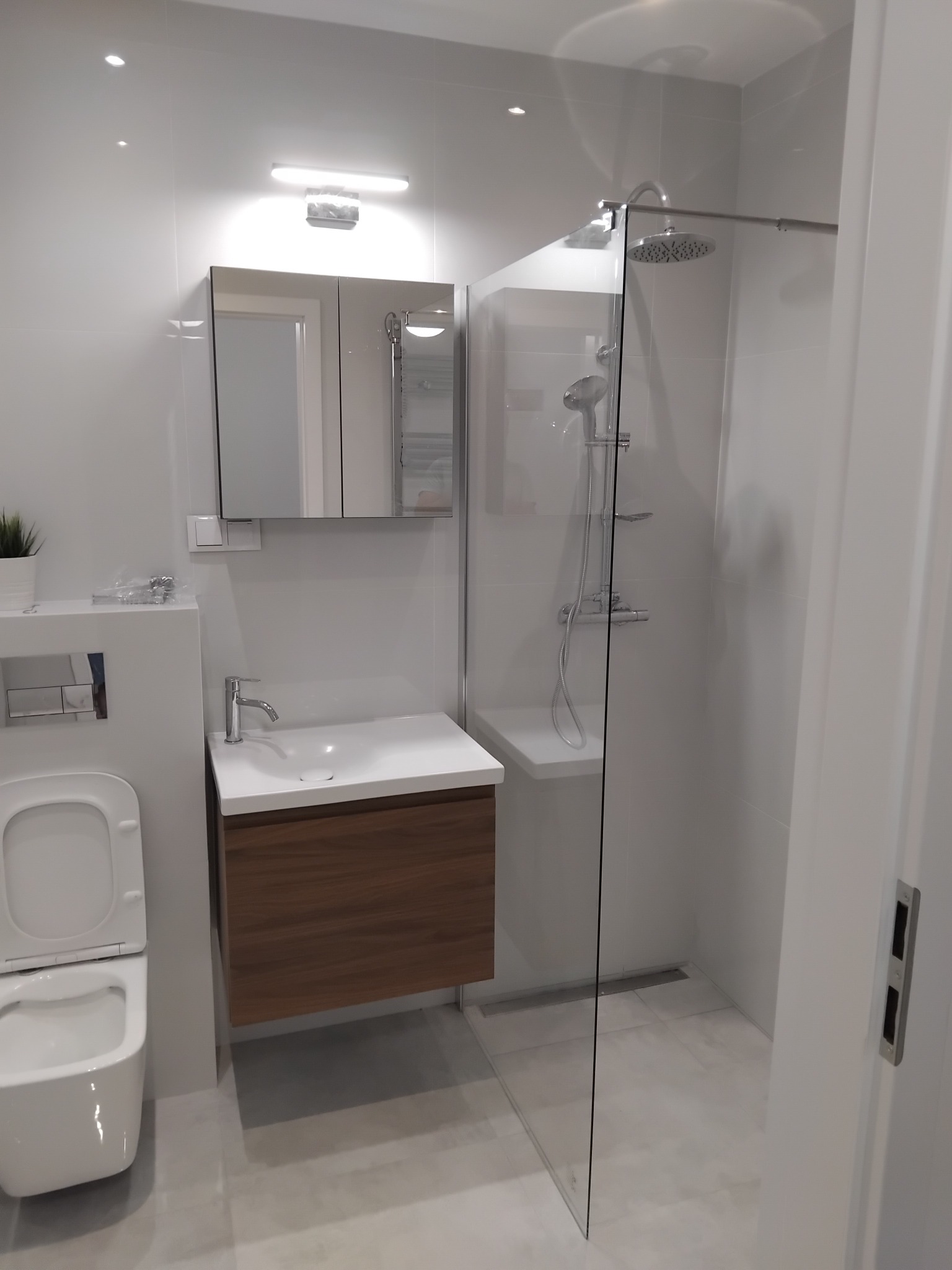 Nowoczesna, jasna łazienka z kabiną prysznicową typu walk-in, umywalką z szafką z drewnianym frontem i podwieszaną toaletą. Minimalistyczny design, szare płytki.