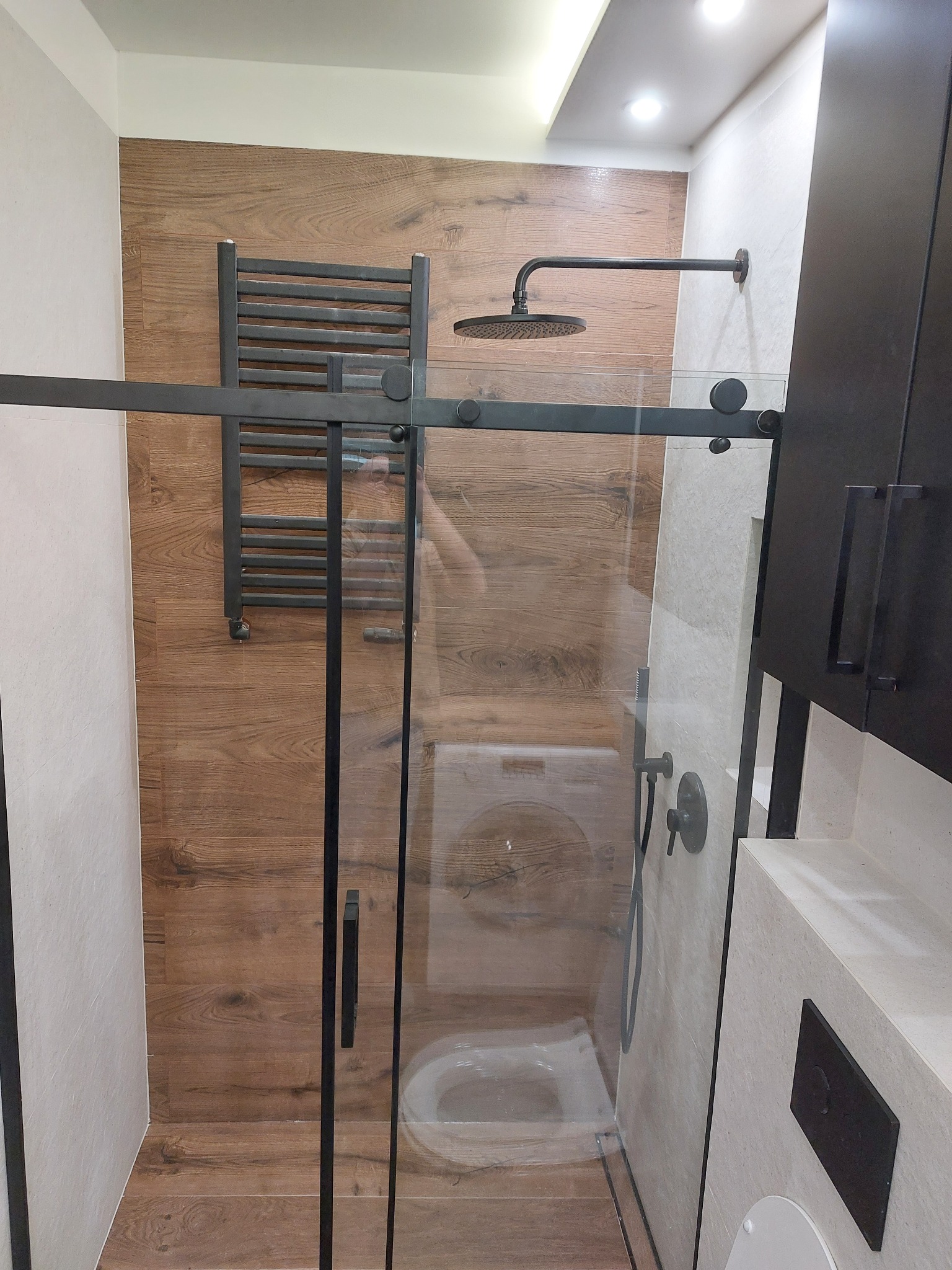 Nowoczesna kabina prysznicowa z czarnymi elementami, drewnopodobne płytki na ścianie, grzejnik drabinkowy. Widoczna pralka w tle, przezroczyste szkło kabiny.