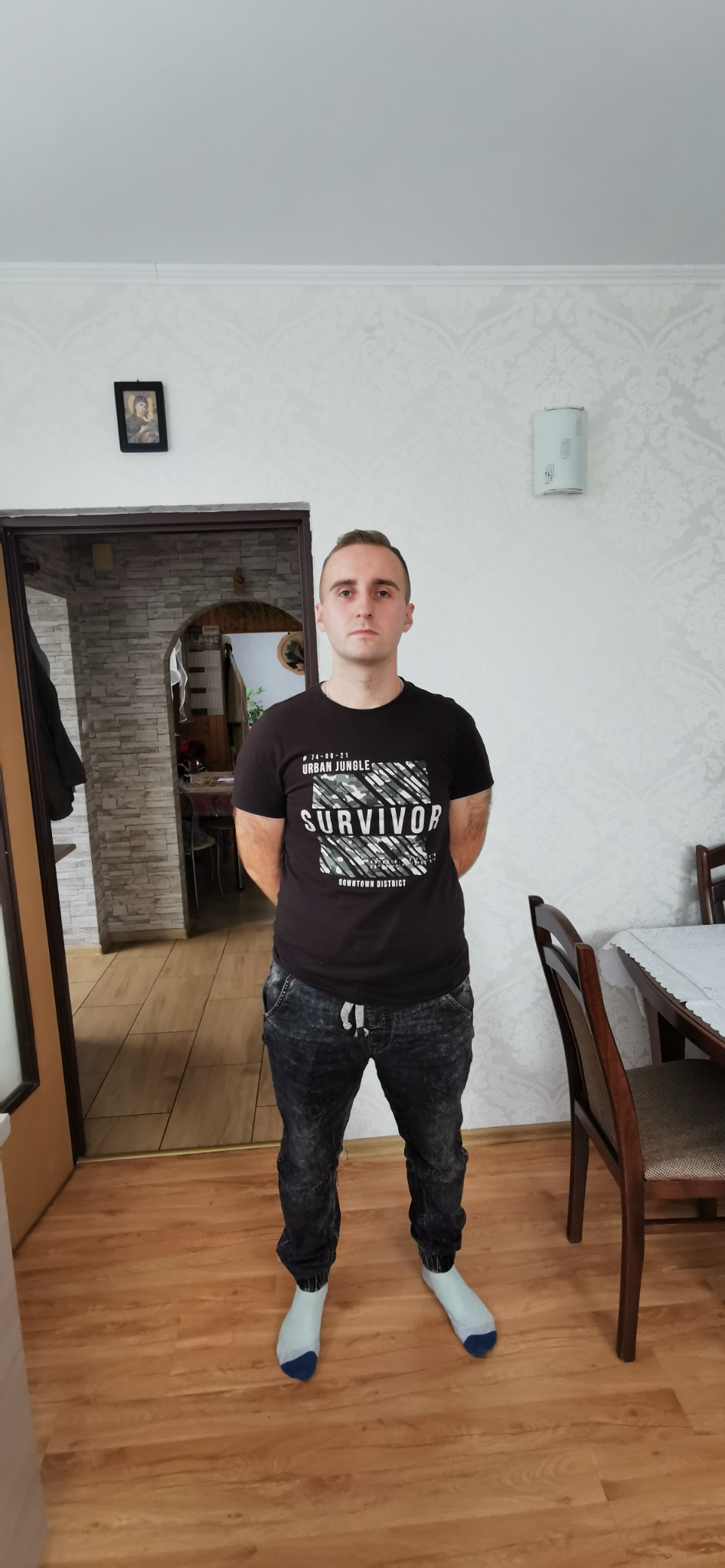 Mężczyzna w czarnej koszulce stoi na tle pokoju z wzorzystą tapetą. W tle widoczne drzwi i fragment jadalni z krzesłem i stołem.