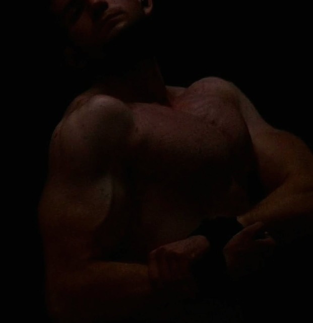 Mężczyzna prezentuje muskulaturę w ciemnym otoczeniu, z mocnym oświetleniem podkreślającym kontury ciała. Widoczne napięcie mięśni ramion i klatki piersiowej.