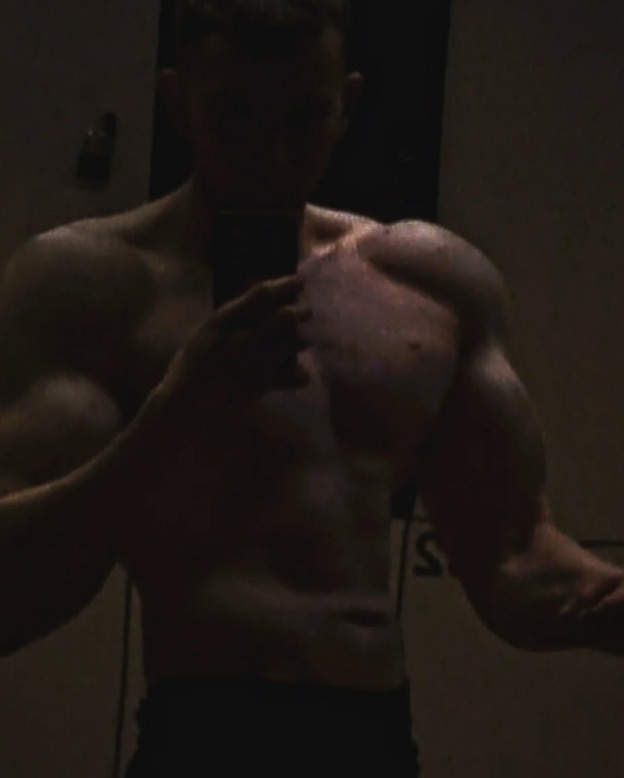 Mężczyzna robi selfie, prezentując swoją umięśnioną sylwetkę w słabym oświetleniu, ukazując efekty treningu. Widoczne zarysy mięśni klatki piersiowej i ramion.