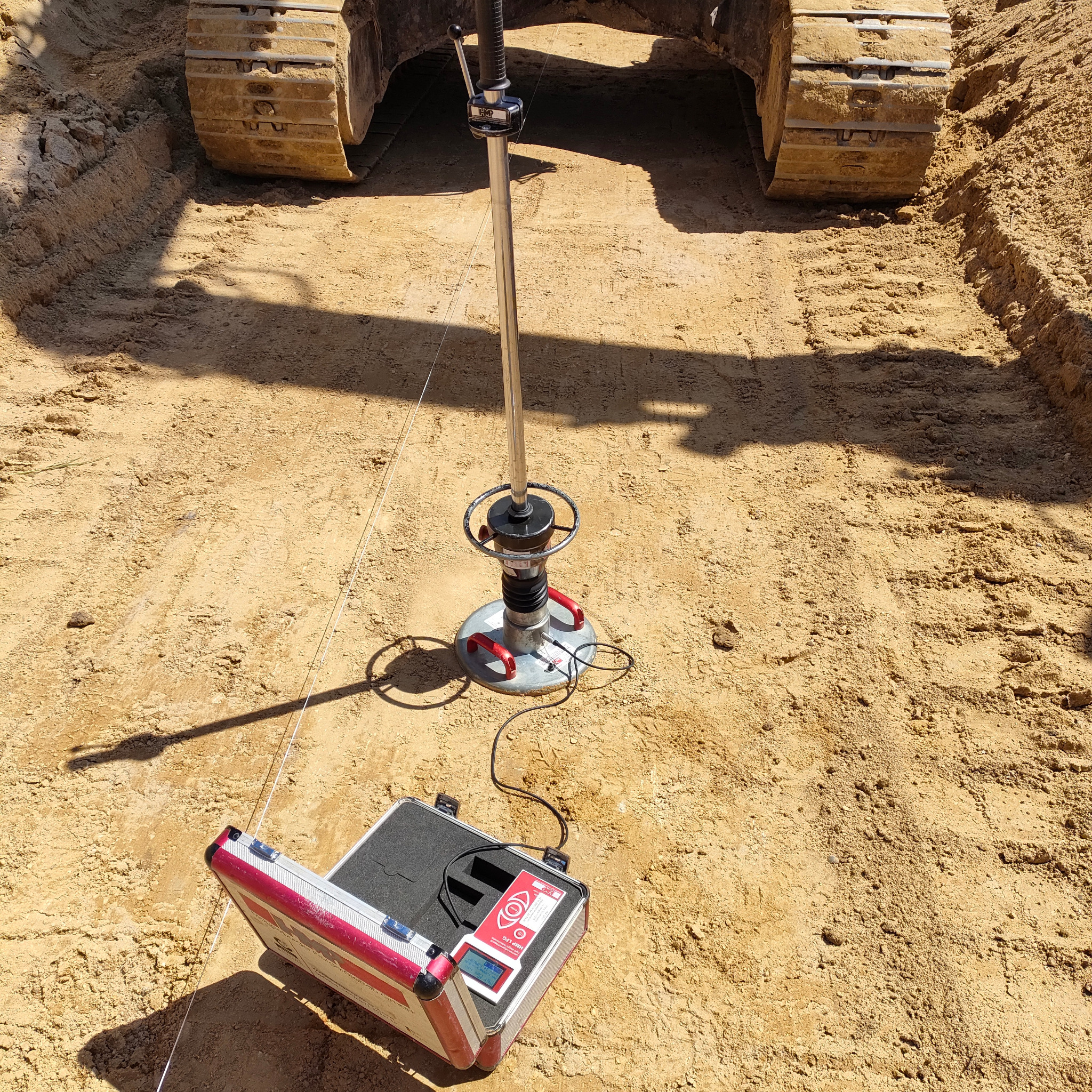 Badanie gruntu w wykopie pod fundamenty z użyciem dynamicznego obciążenia płytą. Widoczne ślady gąsienic koparki i otwarty futerał z urządzeniem pomiarowym.