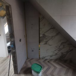 Lara Bud - Łazienka na poddaszu w trakcie wykańczania: ułożone płytki imitujące marmur, podłoga w jodełkę i wiadro z gruntem Knauf Betokontakt. Widoczny fragment surowej ściany.