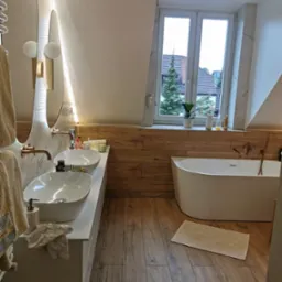 Stylowa łazienka na poddaszu z drewnianą podłogą i panelami ściennymi, owalną wanną, podw&oacute;jną umywalką i oświetlonymi lustrami. Widok na zieleń za oknem.