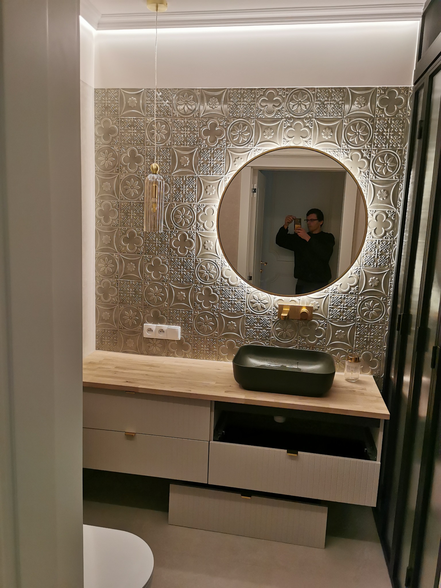 Wykończona łazienka z mozaiką na ścianie, okrągłym lustrem z podświetleniem i czarną umywalką na drewnianym blacie. Widoczne gniazdka i odbicie osoby w lustrze.