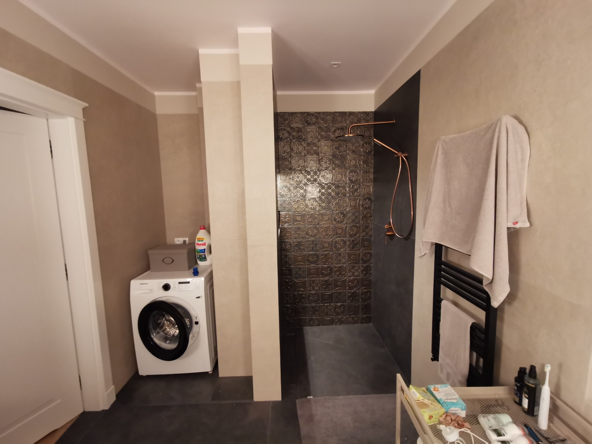 Nowoczesna łazienka z pralką, kabiną prysznicową z dekoracyjną ścianą i miedzianą armaturą. Minimalistyczny design, ciemna podłoga i jasne ściany.