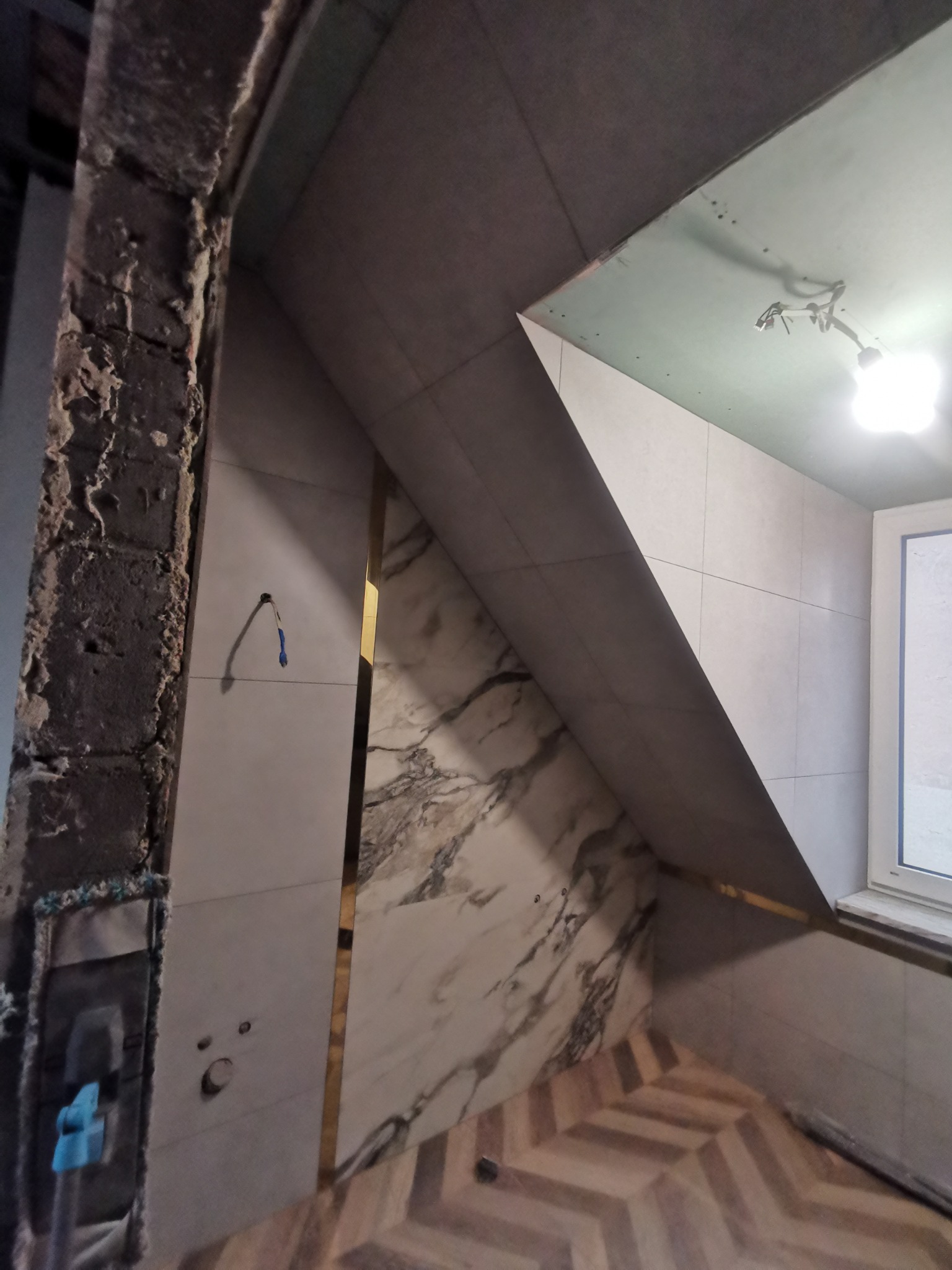 Ukończona łazienka na poddaszu z marmurową ścianą, jasnymi płytkami i drewnianą podłogą w jodełkę. Widoczne niedokończone elementy konstrukcyjne.