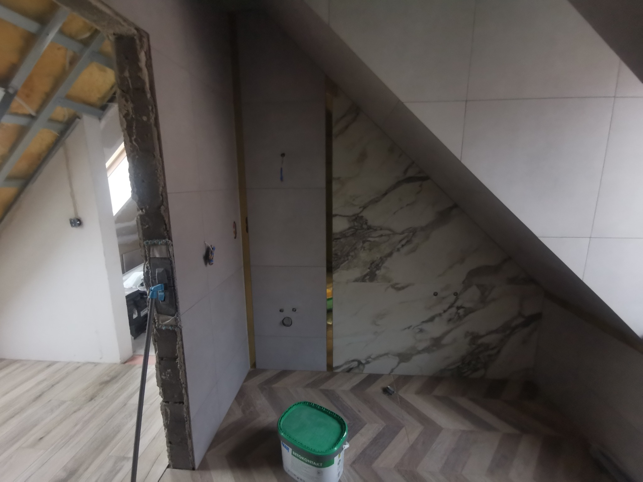 Łazienka na poddaszu w trakcie wykańczania: ułożone płytki imitujące marmur, podłoga w jodełkę i wiadro z gruntem Knauf Betokontakt. Widoczny fragment surowej ściany.