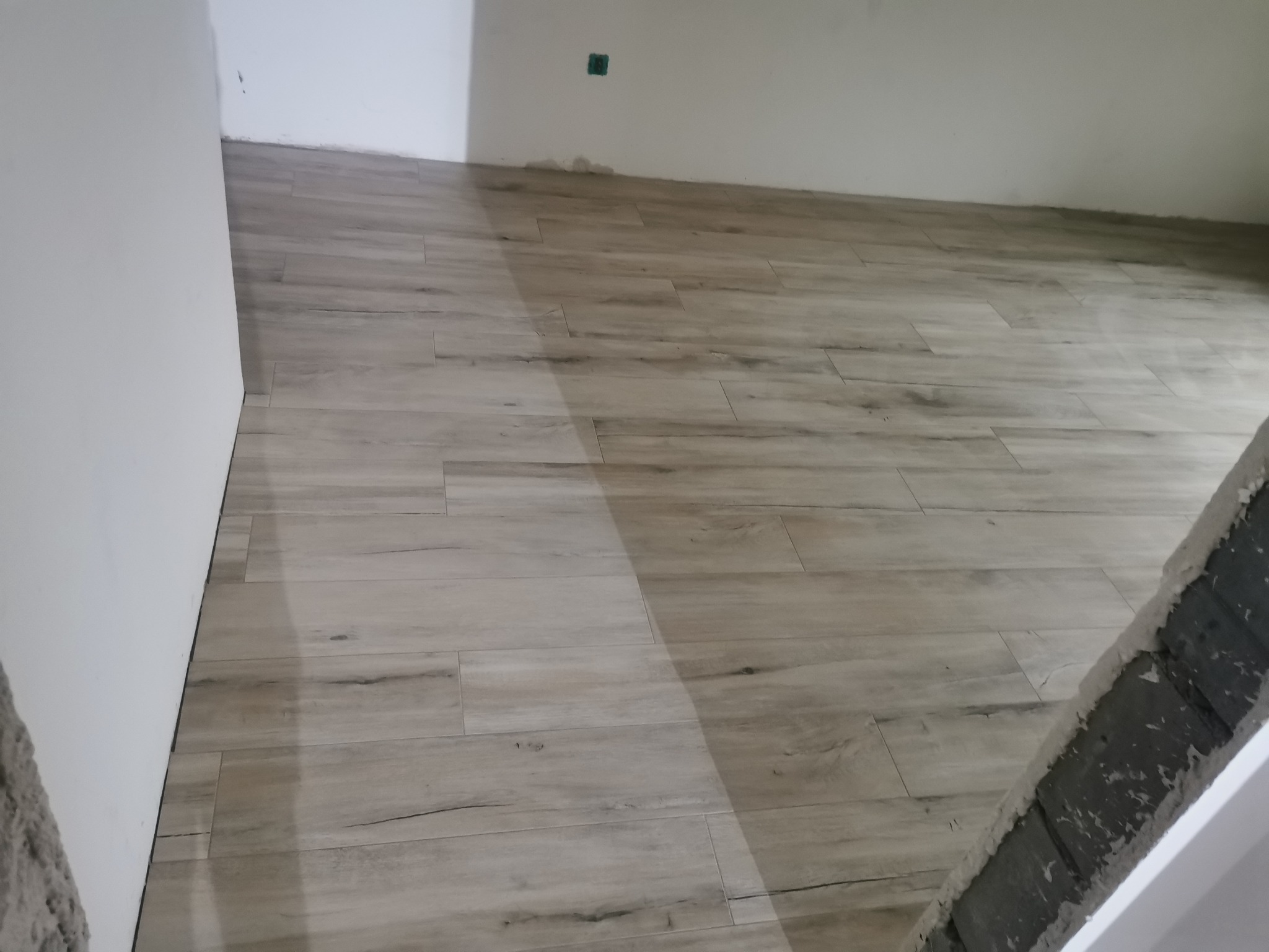Ukończona podłoga z płytek imitujących drewno w jasnym odcieniu, ułożona w nowym pomieszczeniu. Widoczny fragment surowej ściany i narożnika.
