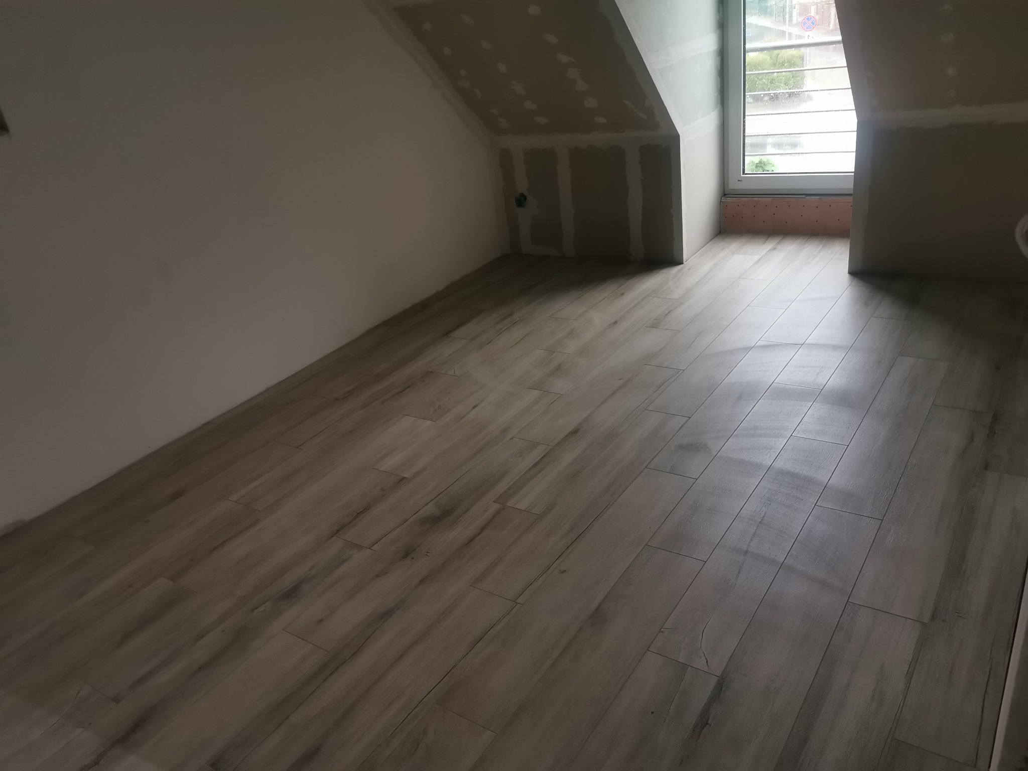 Podłoga z płytek imitujących drewno w pokoju na poddaszu, ściany częściowo wykończone płytami gipsowo-kartonowymi, okno dachowe. Ukończony etap prac.