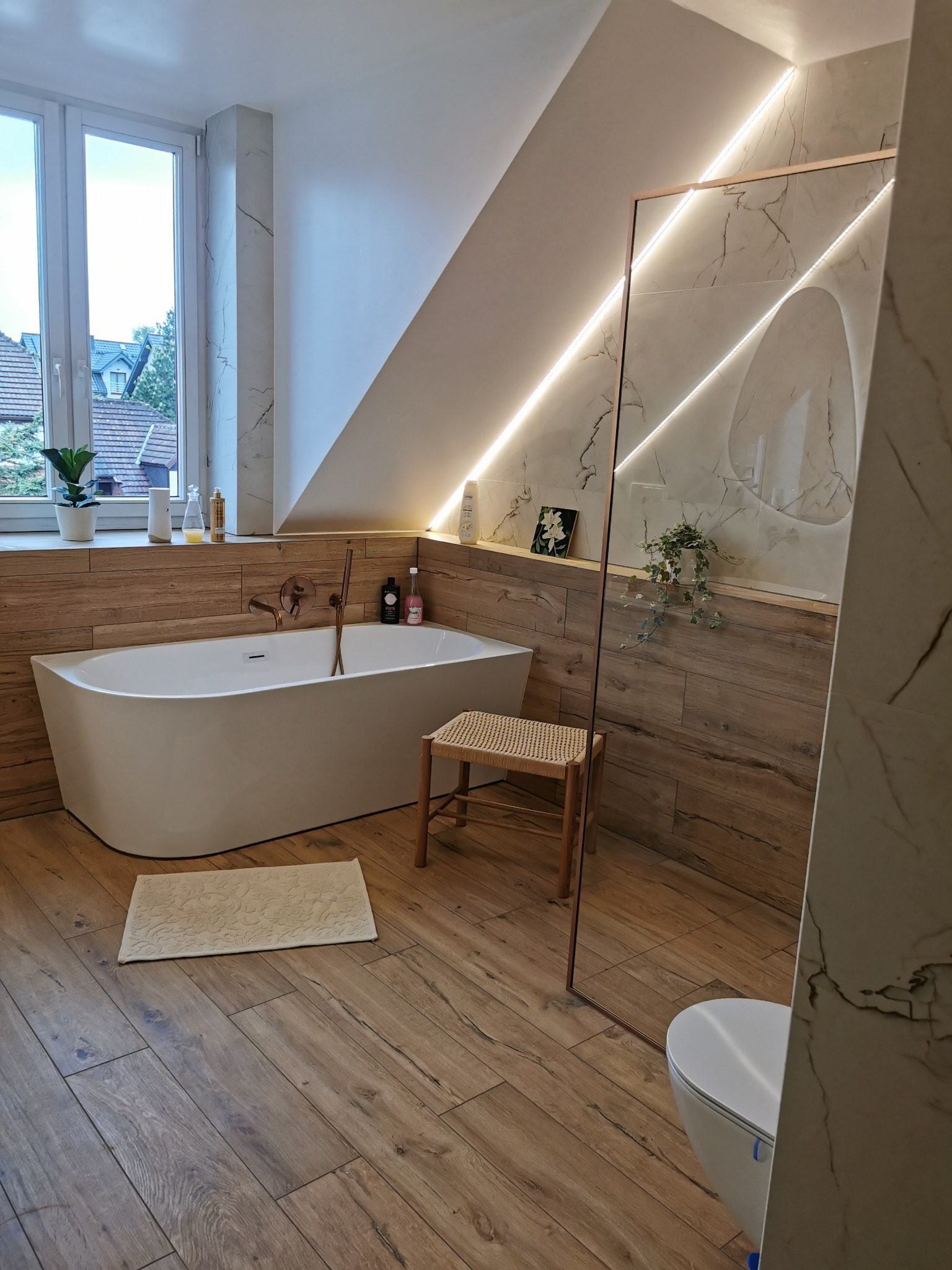 Nowoczesna łazienka na poddaszu z drewnopodobnymi płytkami, wolnostojącą wanną i szklaną kabiną prysznicową z podświetleniem LED.