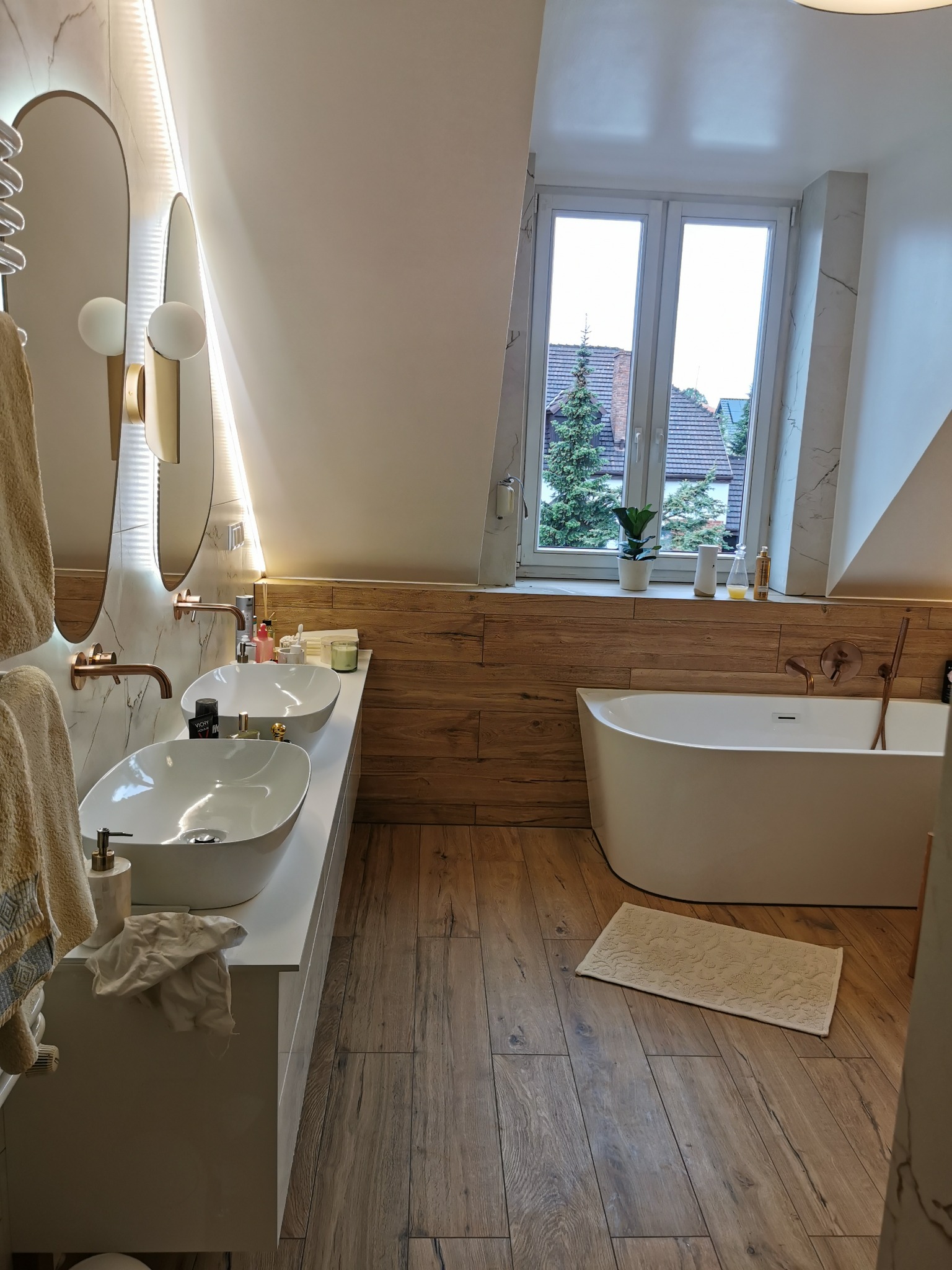 Stylowa łazienka na poddaszu z drewnianą podłogą i panelami ściennymi, owalną wanną, podwójną umywalką i oświetlonymi lustrami. Widok na zieleń za oknem.
