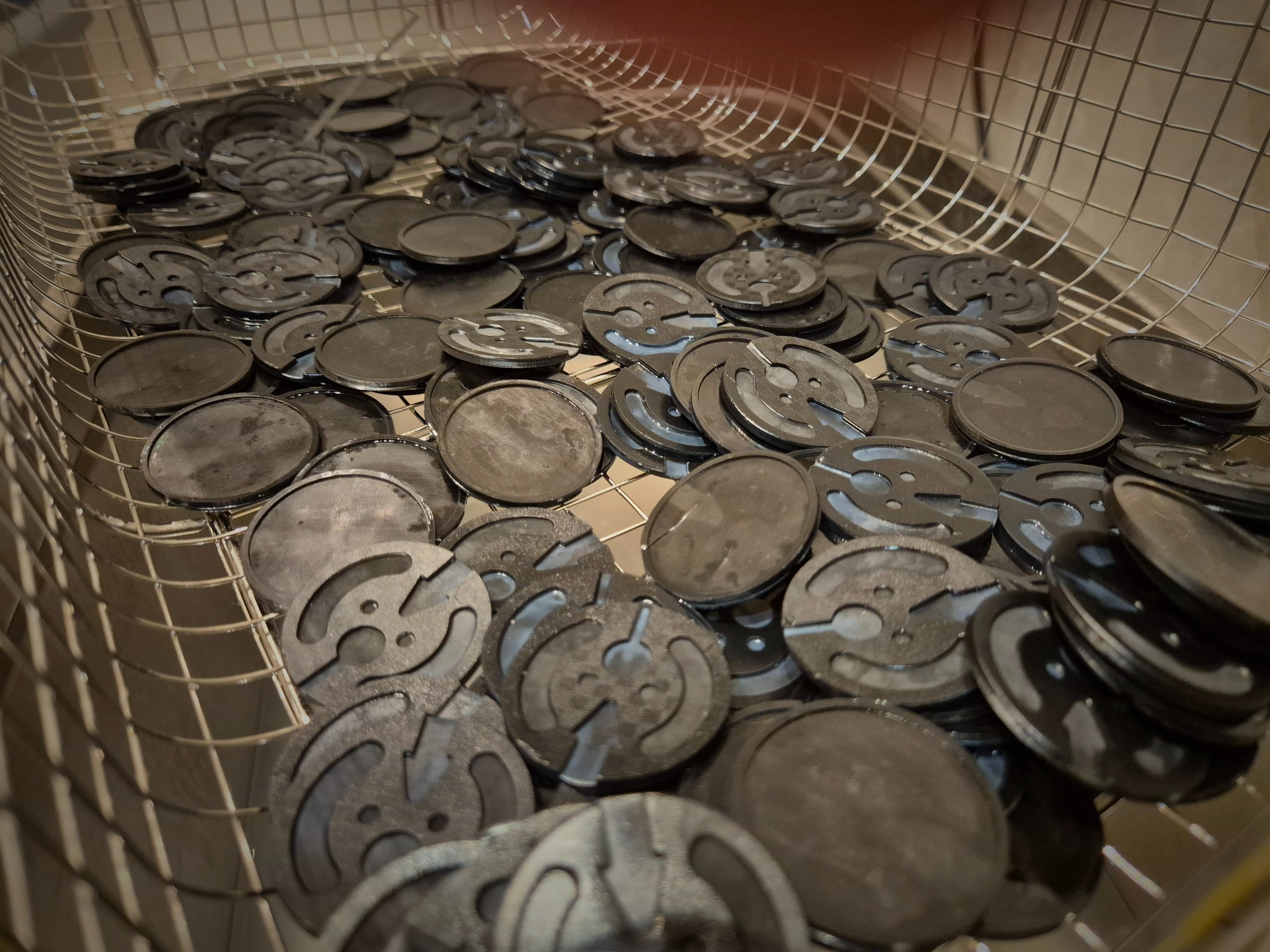 Metalowe, okrągłe elementy z wycięciami w koszu z siatki. Detale obróbki, widoczne krawędzie i otwory. Elementy po procesie czyszczenia lub obróbki wykończeniowej.