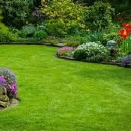 Zadbany, okrągły trawnik otoczony kolorowymi rabatami kwiatowymi i niskim murkiem z kamienia. Bujna roślinność w tle, idealne połączenie zieleni i barw.