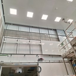 Karol Karasek - Montaż płyt karton-gips na metalowym stelażu w pomieszczeniu z widocznymi elementami wentylacji sufitowej i podnośnikiem. Widok z dołu.