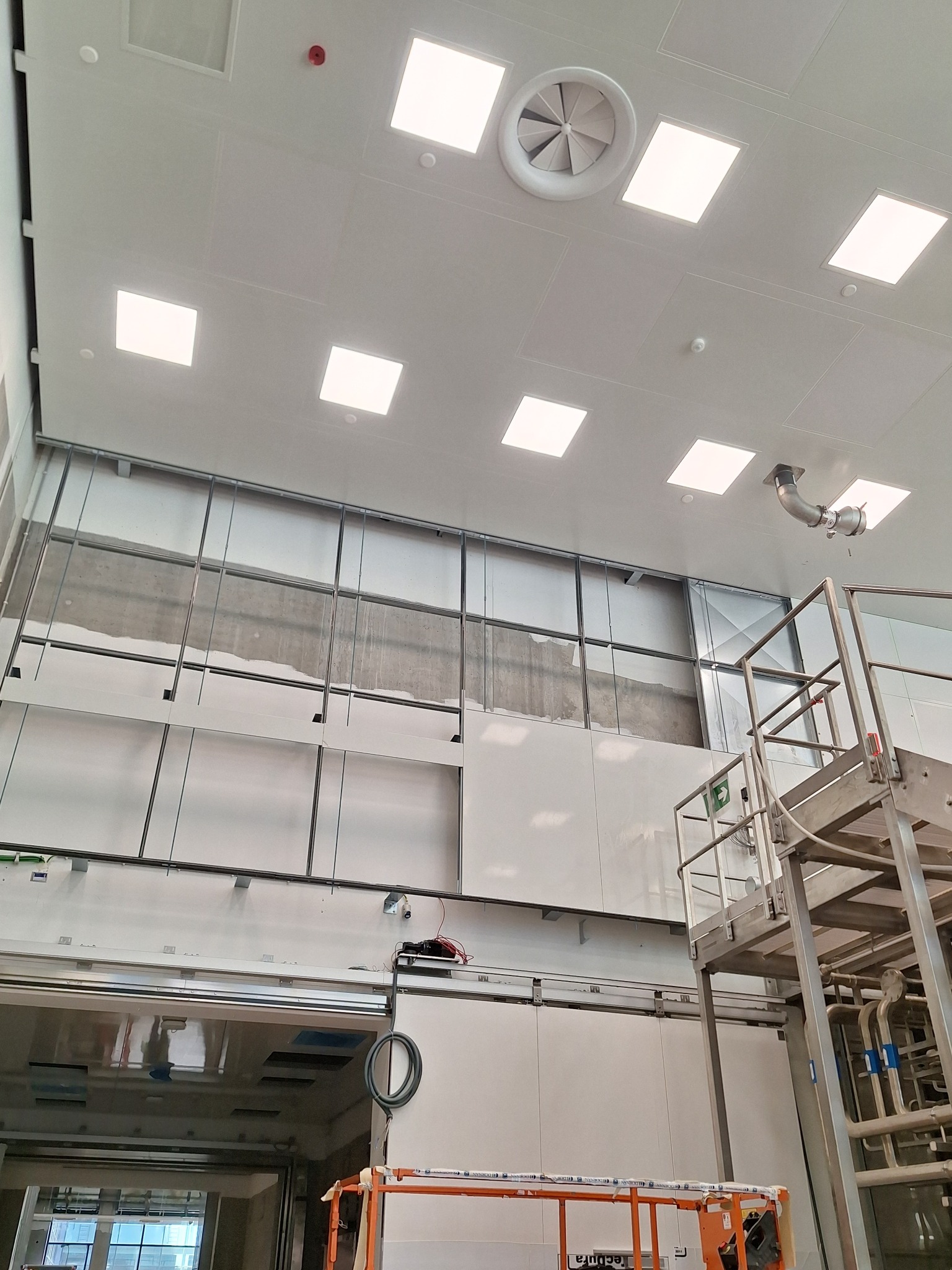 Montaż płyt na ścianie w hali przemysłowej. Widoczne metalowe profile, podnośnik i instalacja wentylacyjna. Oświetlenie sufitowe w kwadratowych panelach.