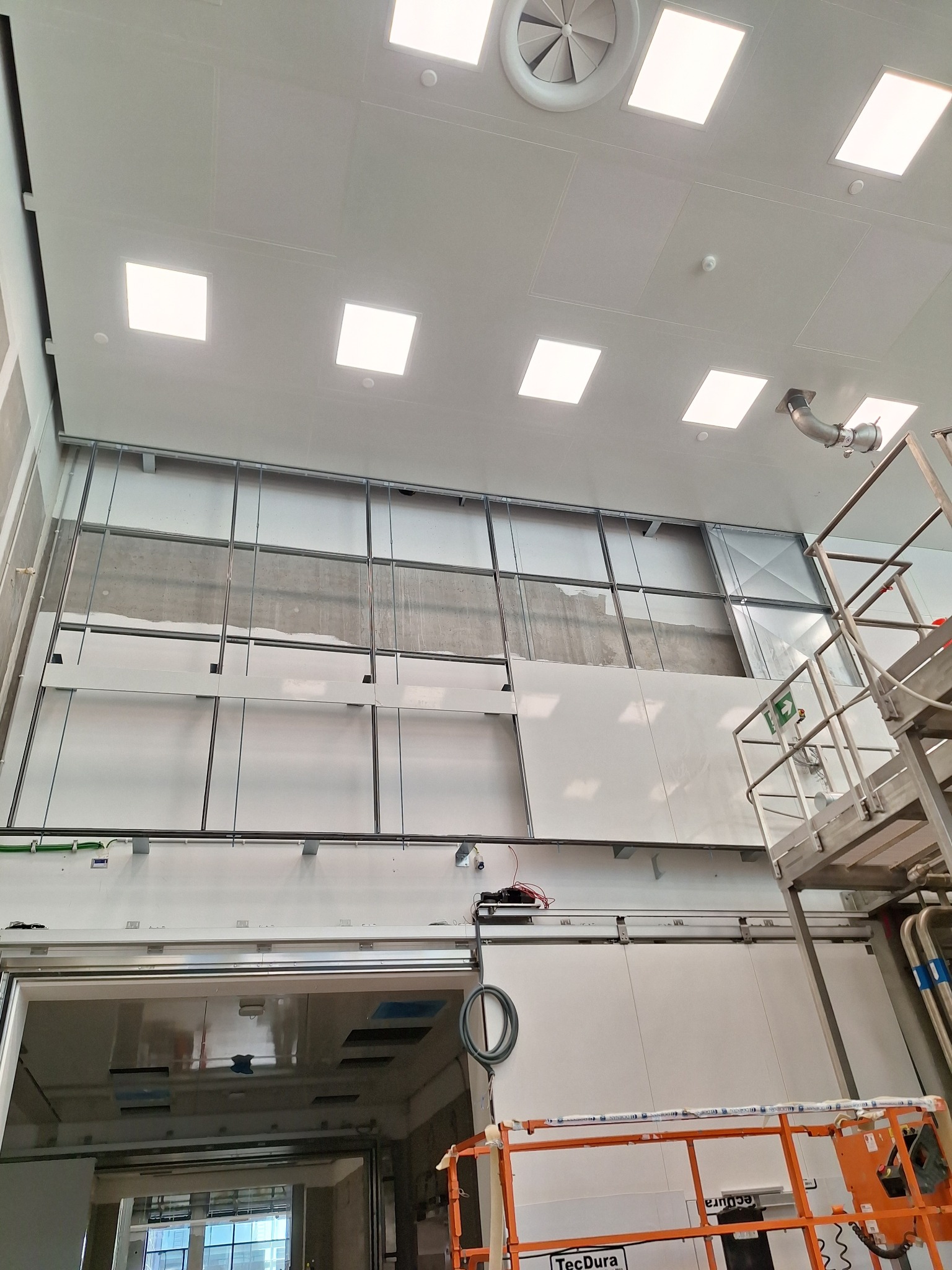 Montaż płyt karton-gips na metalowym stelażu w pomieszczeniu z widocznymi elementami wentylacji sufitowej i podnośnikiem. Widok z dołu.